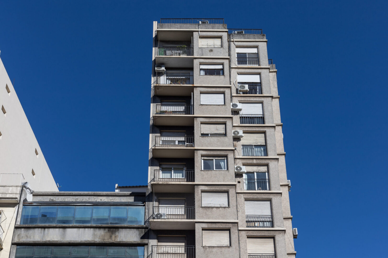 A tall concrete apartment building with balconies and varied window shutters photographed from below against a vibrant blue sky. The weathered brutalist architecture features multiple residential units with air conditioning units and metal railings.